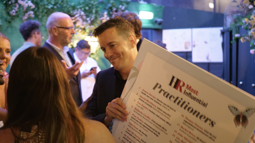 James Devine smiles, holding a printed board listing the top HR practitioners